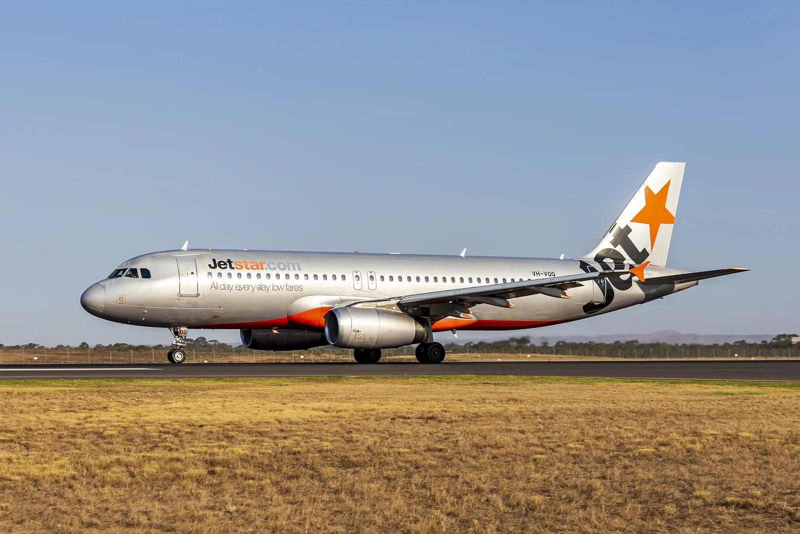 A silver Jetstar Airbus with an orange tail marking on a runway beside dry grass