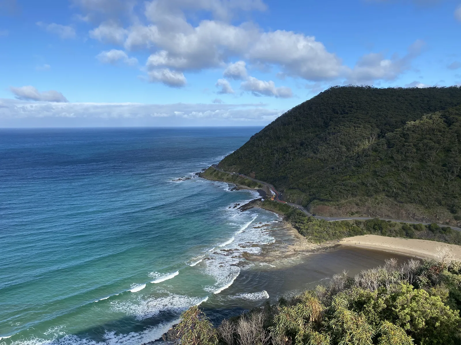 A coastal road beside a sandy beach and a forested headland, with the sea on the left