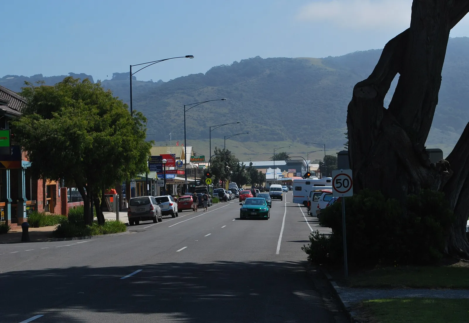 A shopping street with parked cars and green hills behind