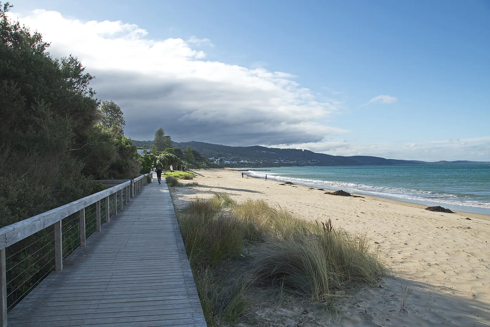 A boardwalk beside dunes and a sandy beach with hills along the coast
