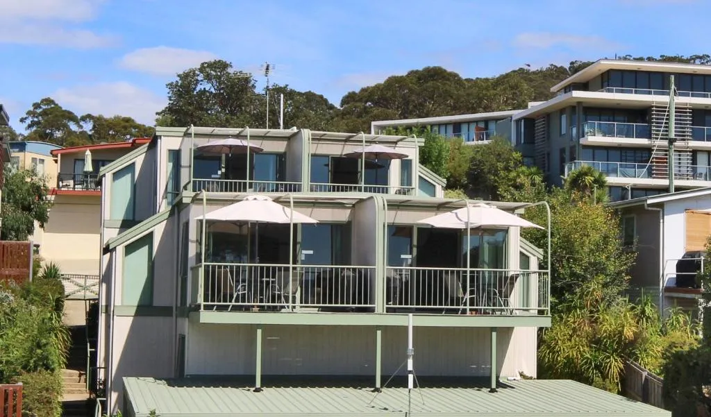 A two-storey building with balconies and white umbrellas, with taller buildings and trees behind