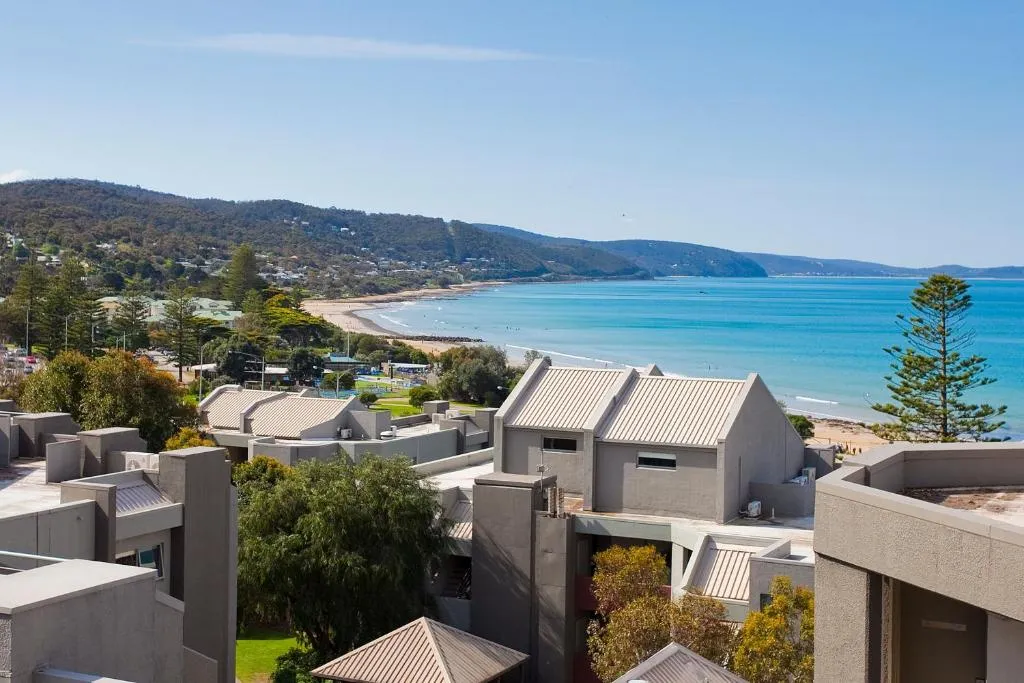 Grey apartment buildings above a curving sandy beach, with blue water and forested hills behind