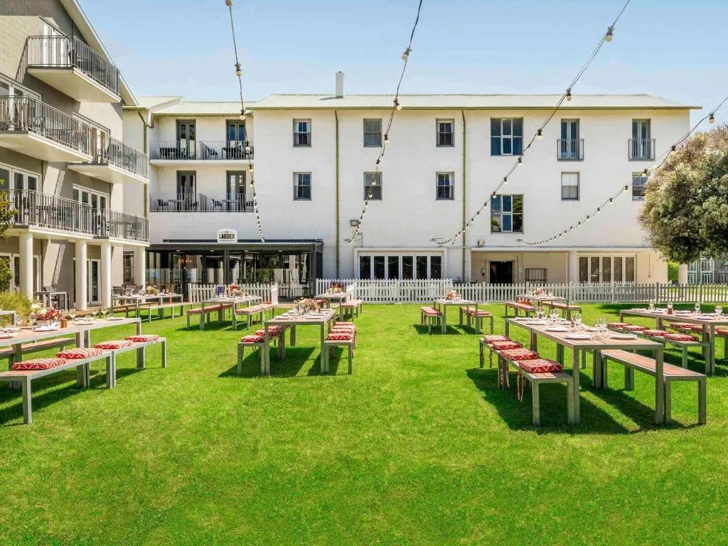 A white multi-storey building behind a lawn set with long outdoor tables and festoon lights